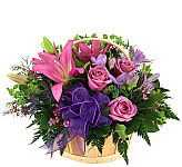 Handled natural woven basket of fresh flowers in pink, lavender and purple with matching ribbon and ample greenery.