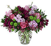 Glass vase arrangement of pink roses, lavender and dark purple stock, green Kermit mums and pink wax flower.
