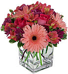 A clear glass cube vase holds pink roses, peach Gerbera daisies, pink alstroemeria and purple daisy poms for any occasion.