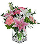 Pink roses, pink and white lilies and pink gerbera are arranged in a cube vase with green berries and foliage.