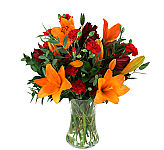 Orange lilies, red alstroemeria, red miniature carnations and greens, all expertly arranged in a round glass vase.