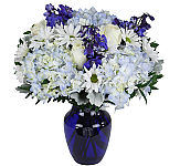 Blue glass vase arrangement of white roses, white daisies, blue hydrangea and blue delphinium.