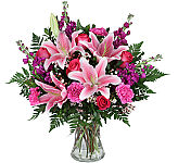 Glass vase arrangement of stargazer lilies, purple stock, hot pink carnations, hot pink roses, wax flower and greens.