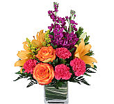 Leaf lined cube vase with orange roses, hot pink carnations, yellow lilies, purple stock and greens.