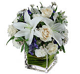 Creme roses, white lilies, carnations and waxflower plus skyblue delphinium in a leaflined glass vase.