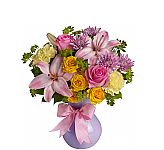 A lavender vase holds yellow spray roses, pink lilies, yellow carnations and lavender mums with added greens and pink ribbon.
