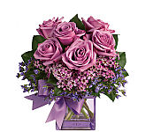 A lavender glass cube vase is expertly arranged with one half dozen lavender roses, lavender waxflower, purple limonium and salal, with a lavender bow.