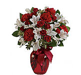 A red glass vase hold red spray roses, red carnations and white alstroemeria with lush greens, tied with a matching red ribbon.